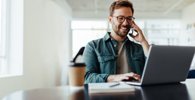 Man smiling and talking on phone while using laptop